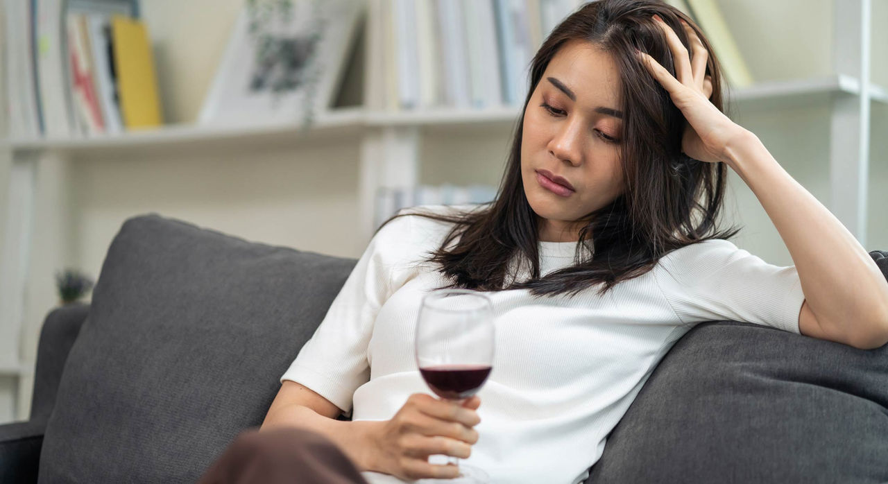 A woman sits on a grey couch and rests her head on one hand while holding and staring at a wine glass filled with some red wine