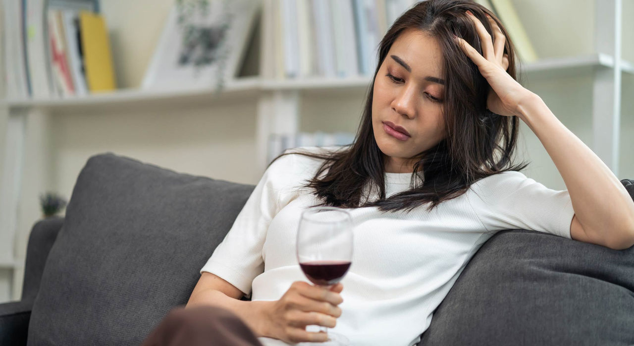 Alcohol and Heart Disease A woman sits on a grey couch and rests her head on one hand while holding and staring at a wine glass filled with some red wine