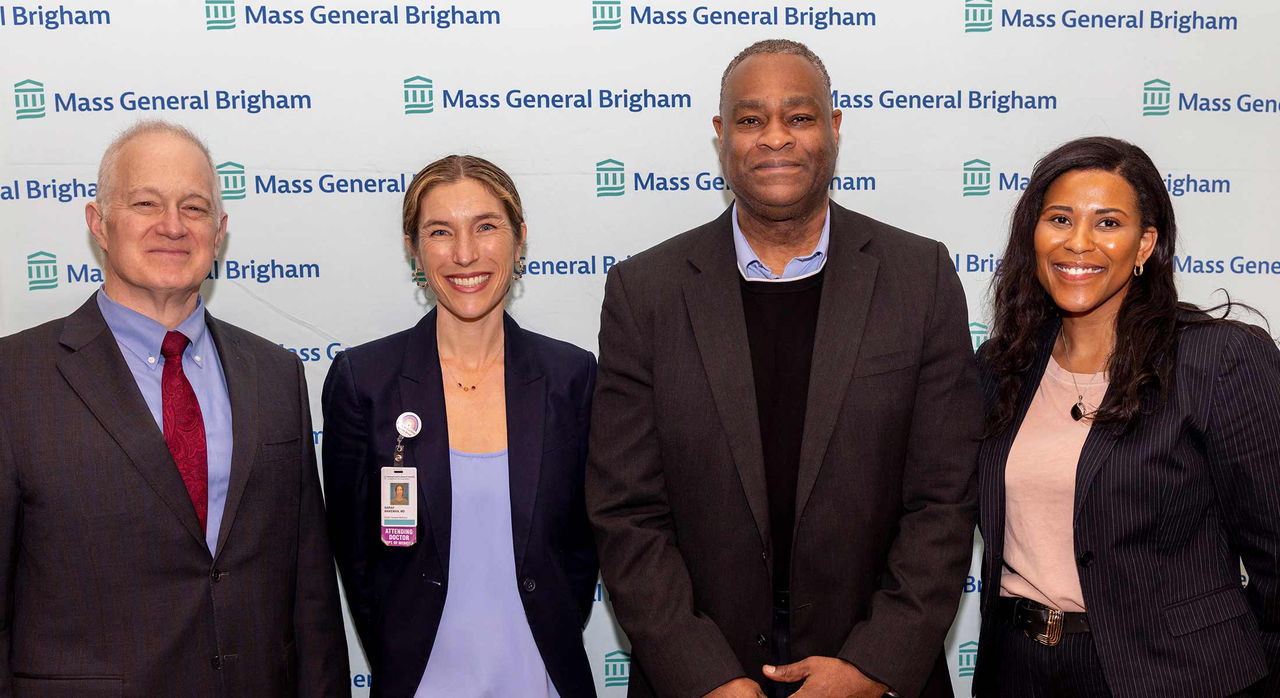 Four individuals in professional clothing standing and smiling together in front of a Mass General Brigham step-and-repeat background