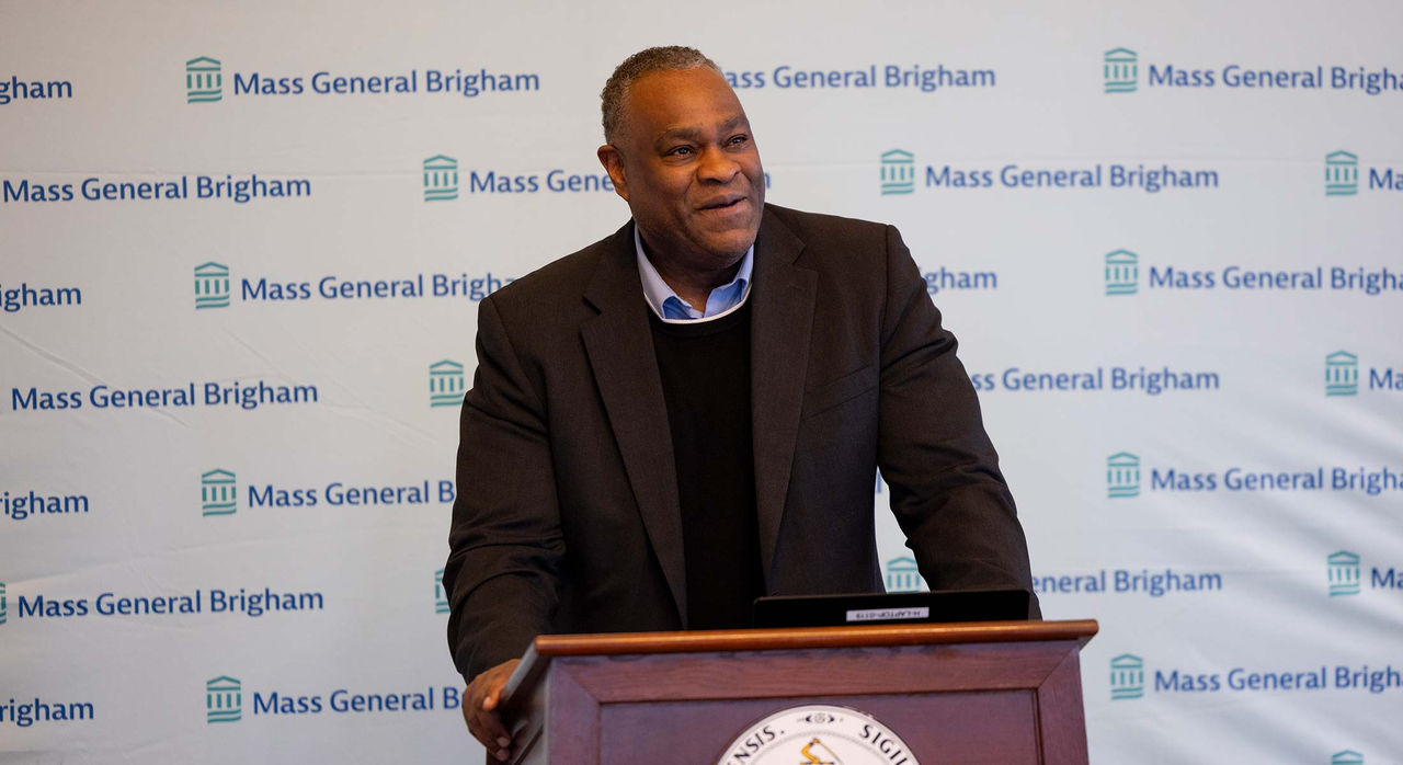 A man in a shirt, sweater, and blazer speaks from a podium with a white background featuring the Mass General Brigham logo behind him