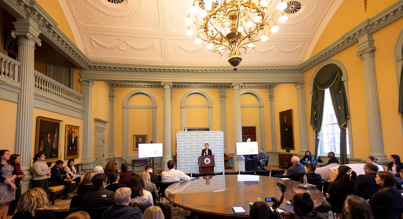 A person speaking at a podium in a room full of people at the Massachusetts State House