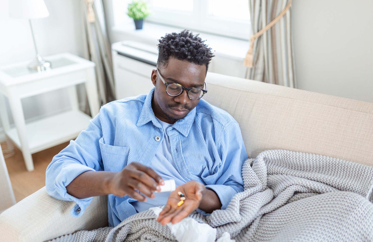 A Black man wearing glasses, a blue undershirt and overshirt, is sitting on the couch and pouring out a few tablets from his right hand to his left palm