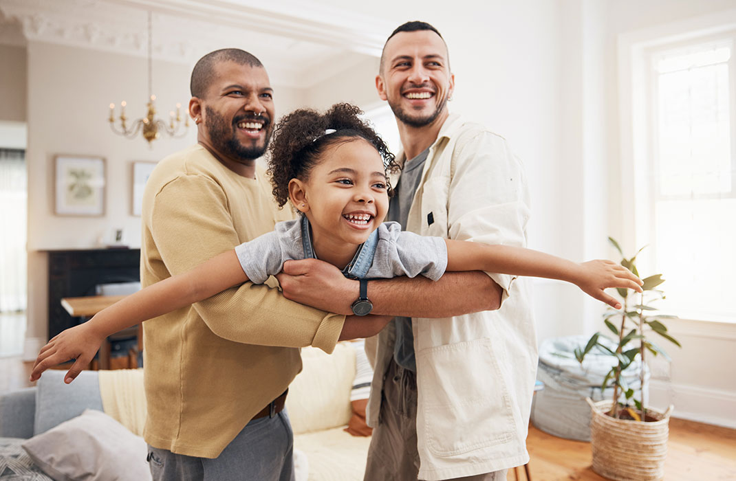 Two Black men hold up their smiling young daughter, who spreads her arms like she's an airplane.