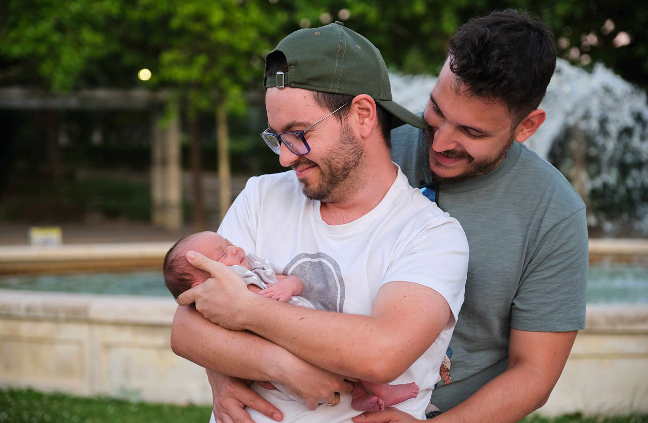 Two young  white men look adoringly at the baby that one of the men holds.
