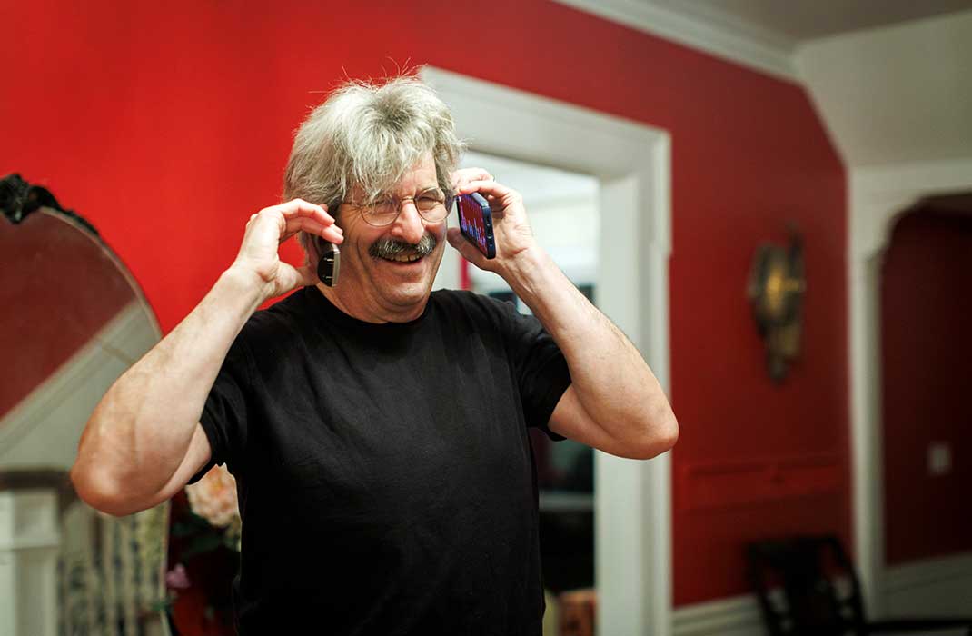 An individual smiling and holding two mobile phones up to is ears.