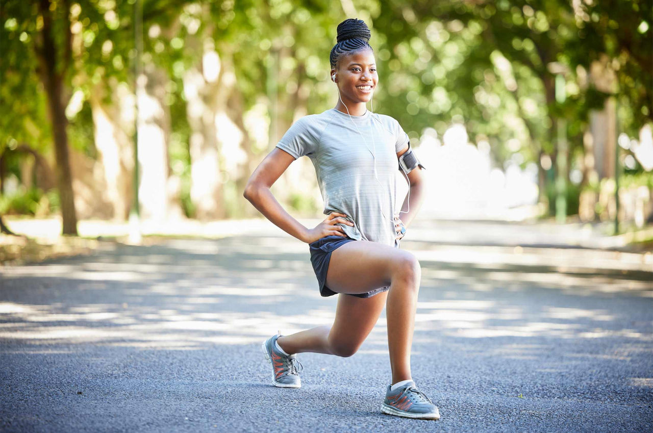 A woman athlete wearing a grey top, black shorts, and running shoes does hip mobility exercises outdoors