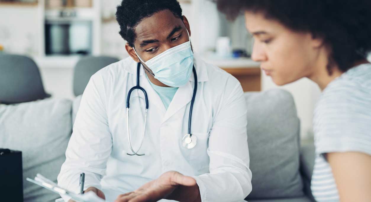 A masked doctor discusses a clipboard of information with a patient.