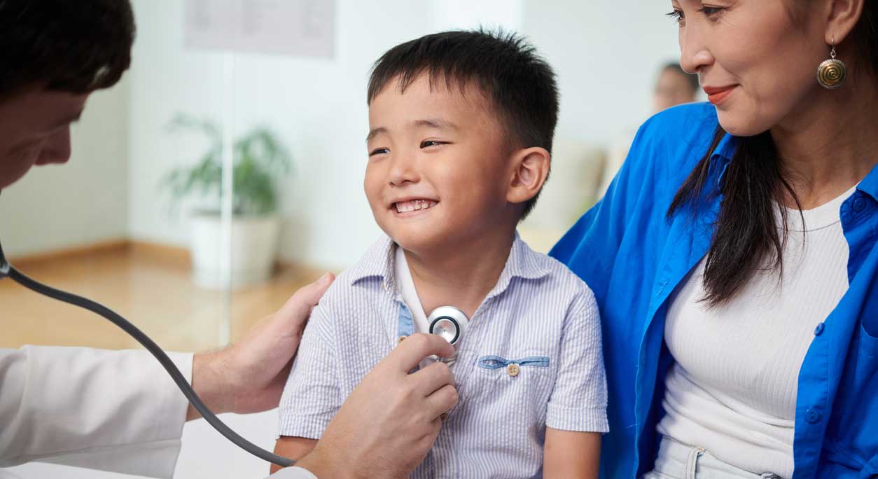 A doctor listens to a smiling little boy's heart while his mother sits beside him.