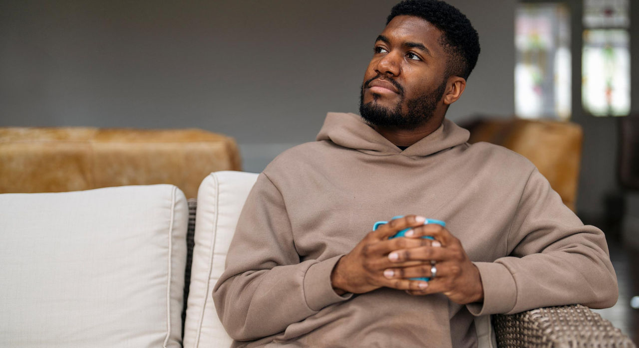 A Black man, wearing a brown hoodie, smiles and looks off to his right side as he holds a blue mug while sitting on a couch