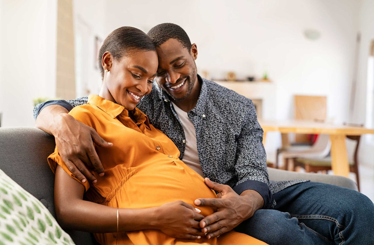 A smiling couple sits on their couch, cradling the woman's pregnant stomach.
