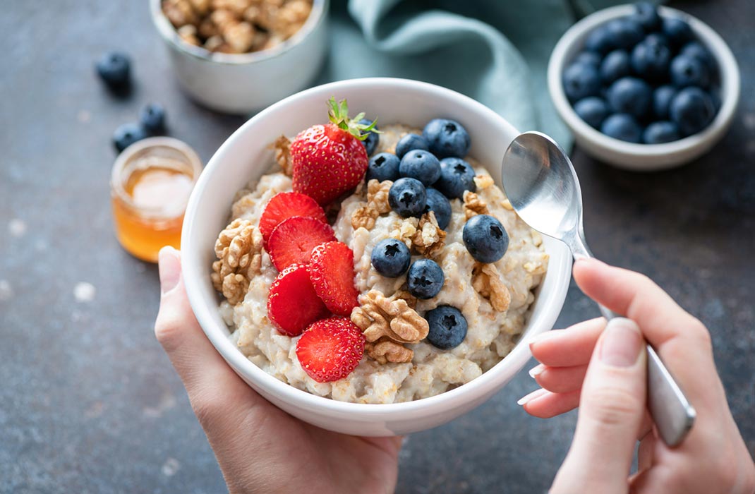 A white bowl of oats, walnuts, strawberries, and blueberries held by a person using both hands with a spoon in the right hand