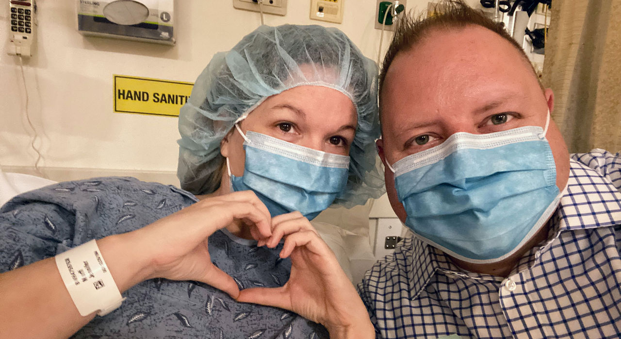 Jenn and her husband at the hospital. Jennifer is gowned, masked, and wearing a hairnet, and she makes the shape of a heart with her hands.
