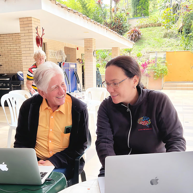 Two researchers, an older man and a woman, on laptops in a sunny courtyard.