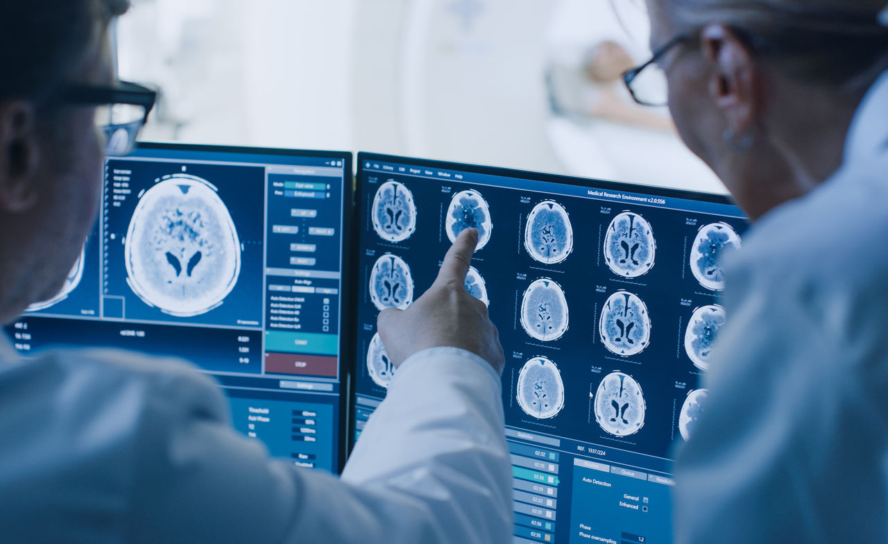 In Control Room Doctor and Radiologist Discuss Diagnosis while Watching Procedure and Monitors Showing Brain Scans Results, In the Background Patient Undergoes MRI or CT Scan Procedure.