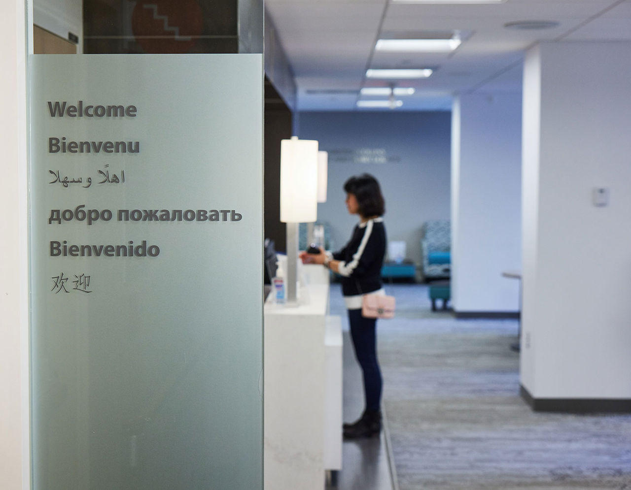 A welcome sign in many languages, with the office visible through the glass wall.